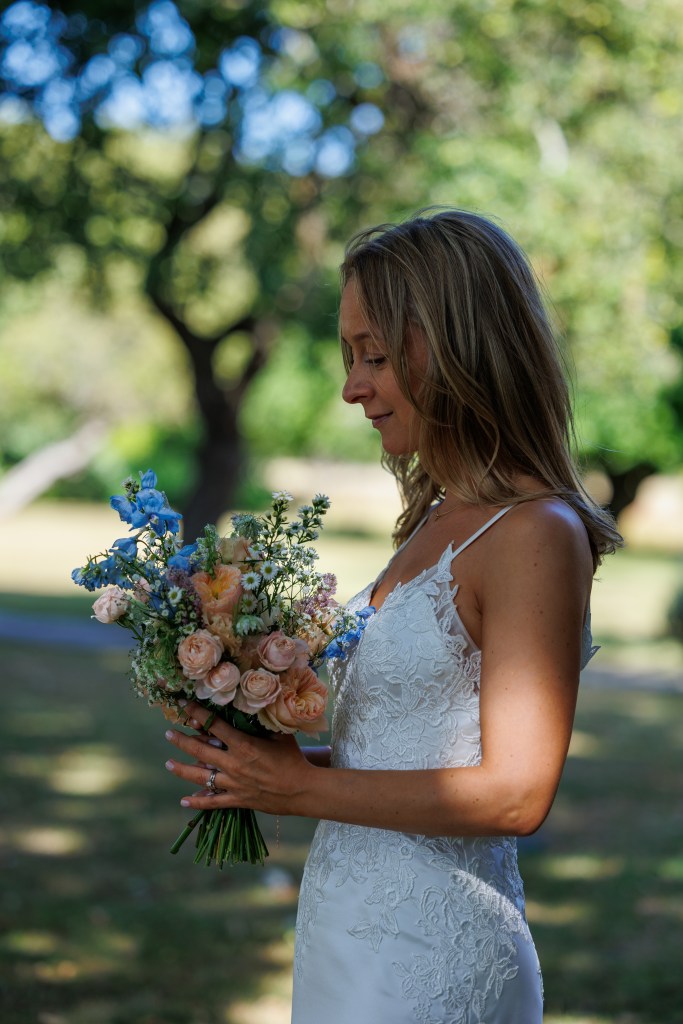 Bride in a lace wedding dress standing in soft natural light at Bailiffscourt Hotel and Spa in Climping, West Sussex, holding and admiring a bouquet of pastel roses and blue flowers. Surrounded by blurred greenery and trees, this calm, intimate moment highlights natural light and fine detail, captured in a relaxed documentary style by a Sussex wedding photographer.