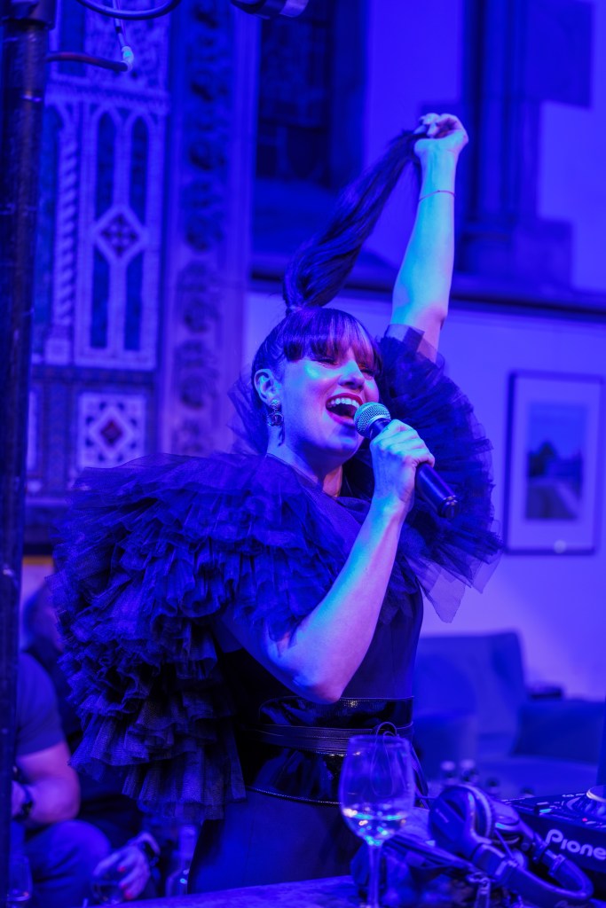 Tara McDonald performing live at a Christmas party at Graylingwell Chapel in Chichester, singing into a microphone under vibrant blue stage lighting. The singer wears a dramatic black ruffled outfit and lifts her hair mid-performance, creating a high-energy moment captured during a Stylishouse event by a Chichester event photographer.