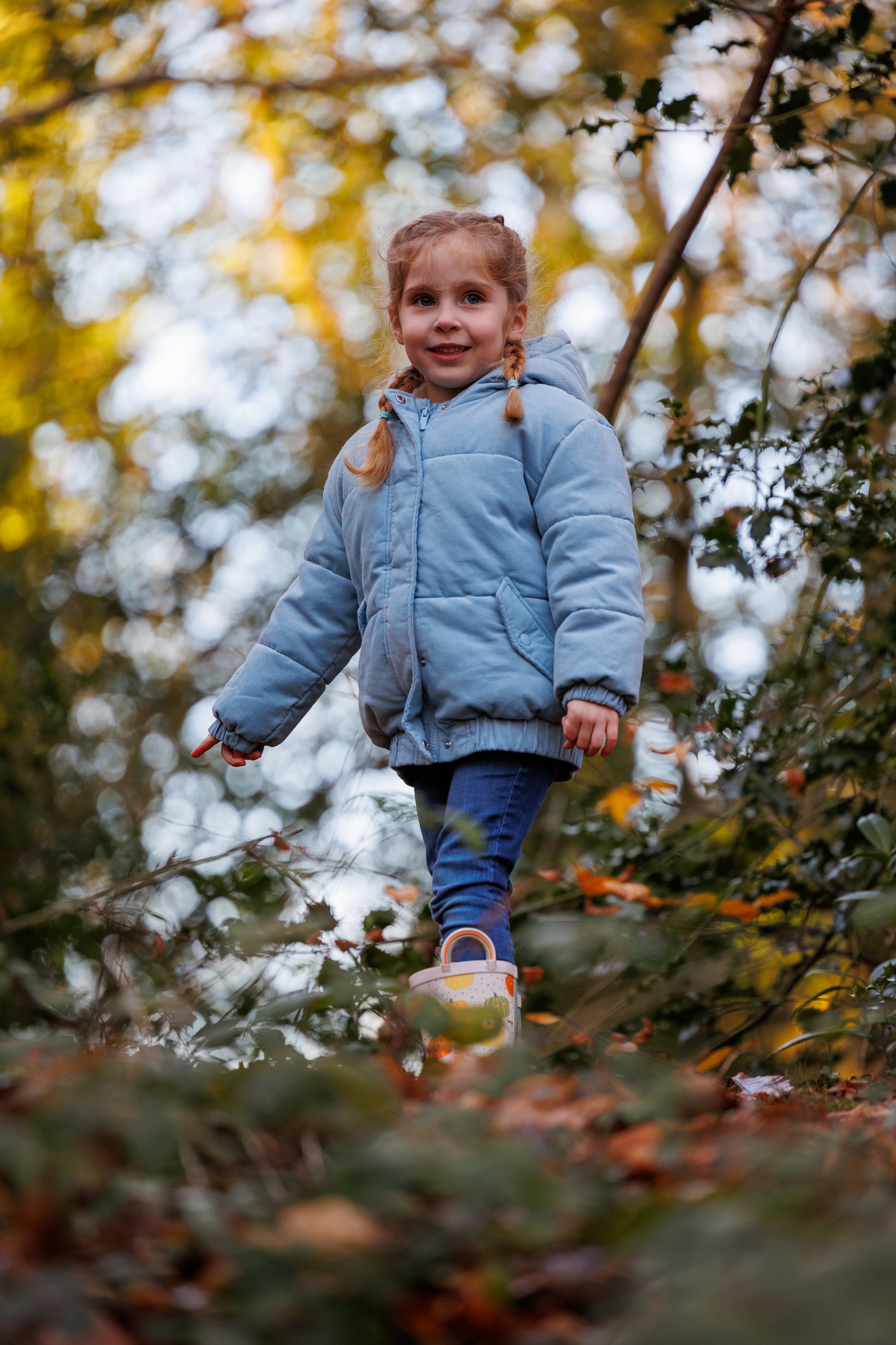 Young child standing among woodland undergrowth at Brandy Hole Copse in Chichester during an outdoor portrait photography session. Wearing a blue jacket and surrounded by autumn leaves and soft natural light, the image captures a candid, playful moment in a relaxed family portrait style by a Chichester portrait photographer.