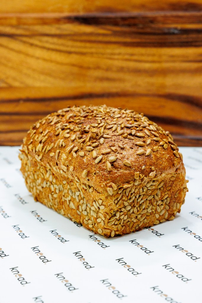 Seeded artisan loaf with a golden crust and mixed seeds on top, photographed on branded Knead bakery paper against a warm wooden background, highlighting texture and fresh bakery quality