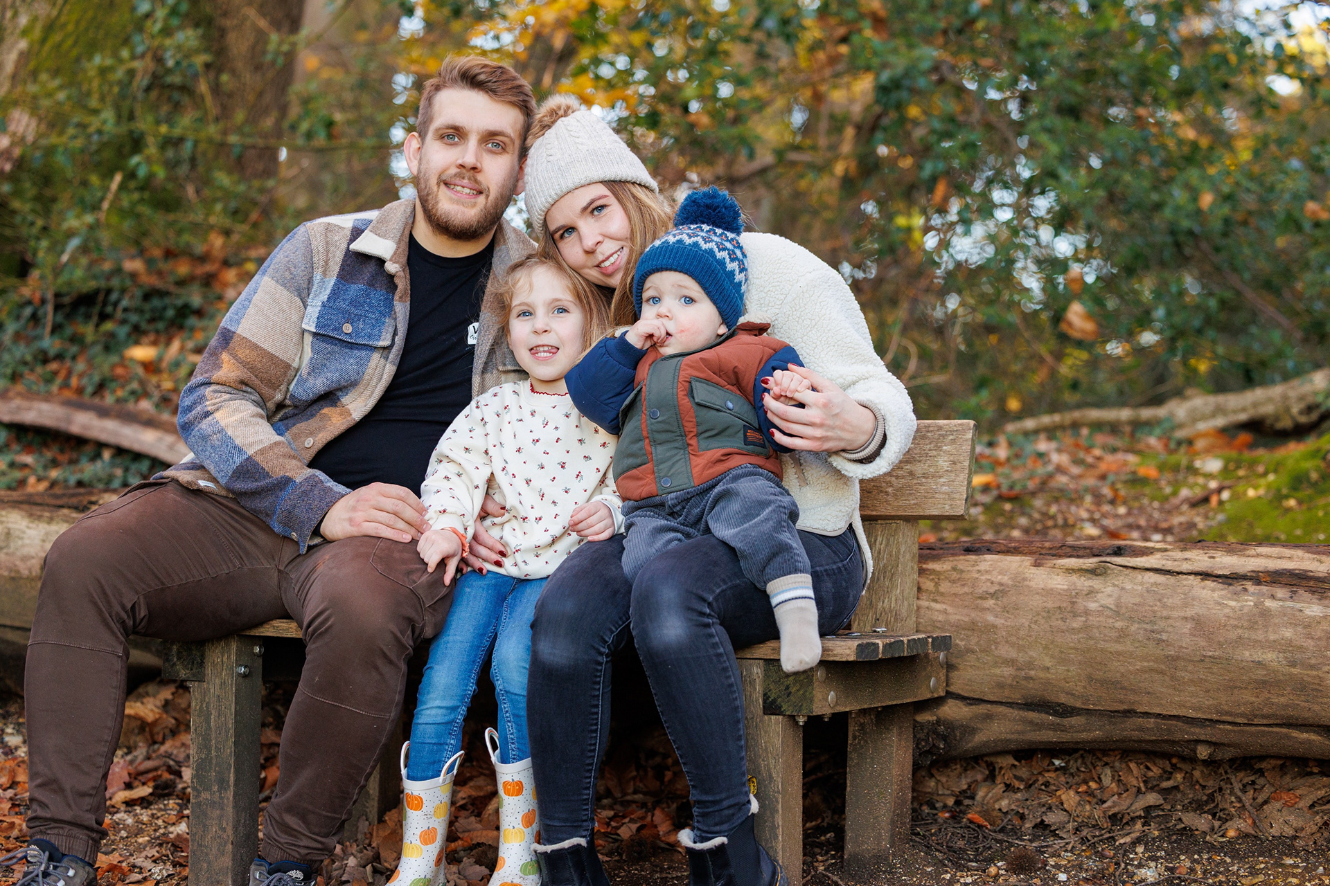 Family of four sitting together on a wooden bench in an autumn woodland during an outdoor portrait session.