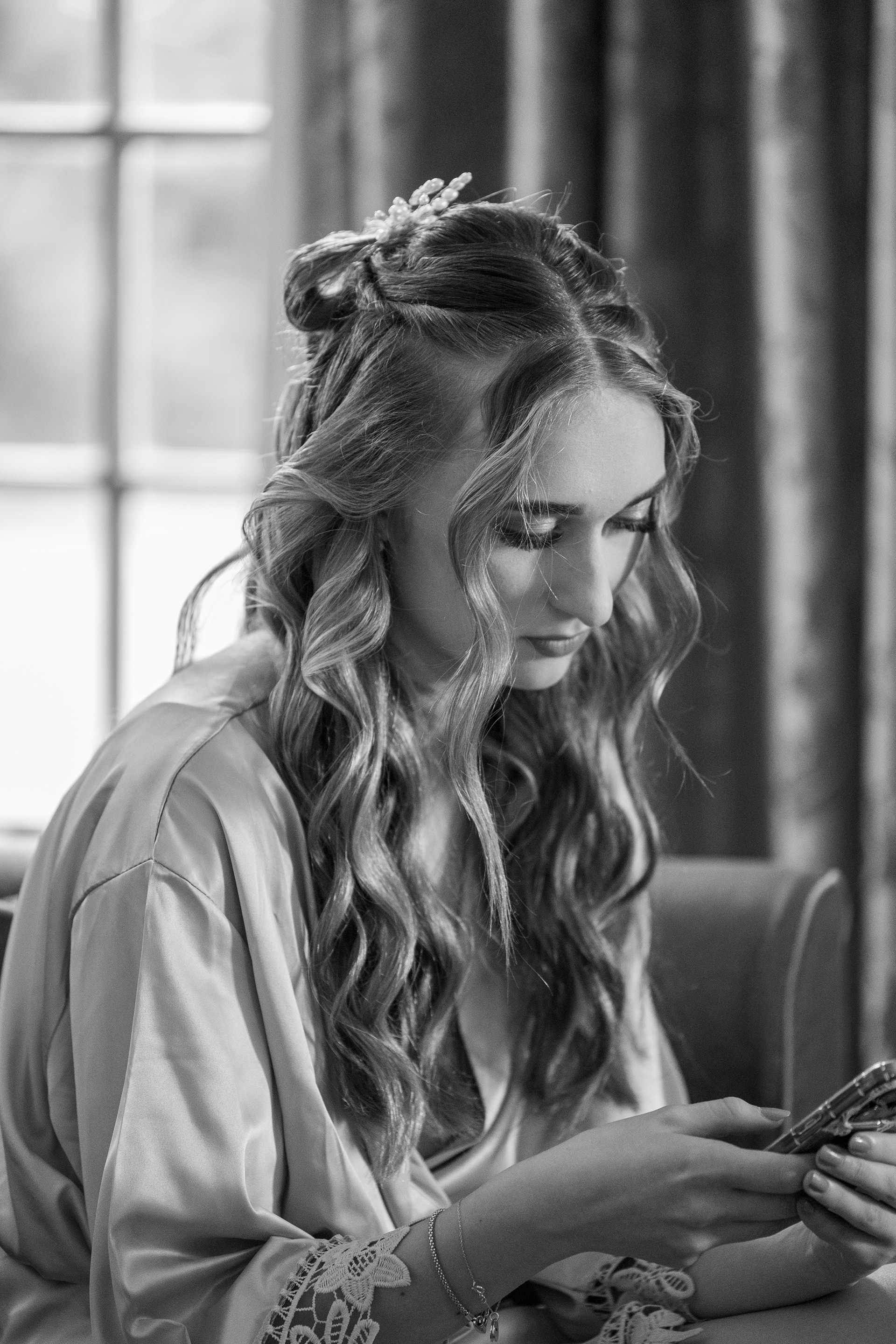 Black and white portrait of a bride during wedding preparations, seated indoors in a satin robe while looking at her phone beside a window.