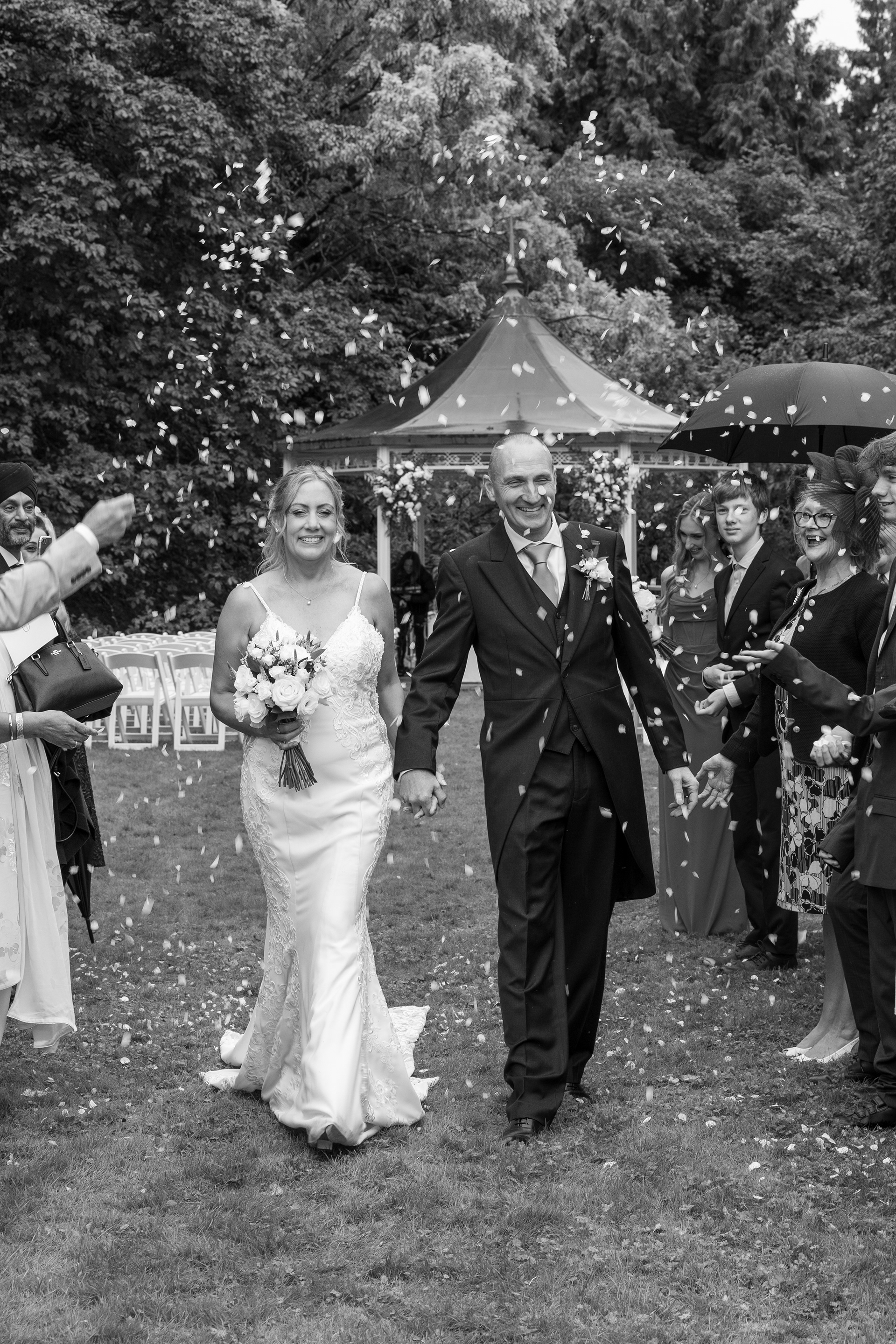 Black and white photograph of a bride and groom walking through confetti thrown by guests after an outdoor wedding ceremony.