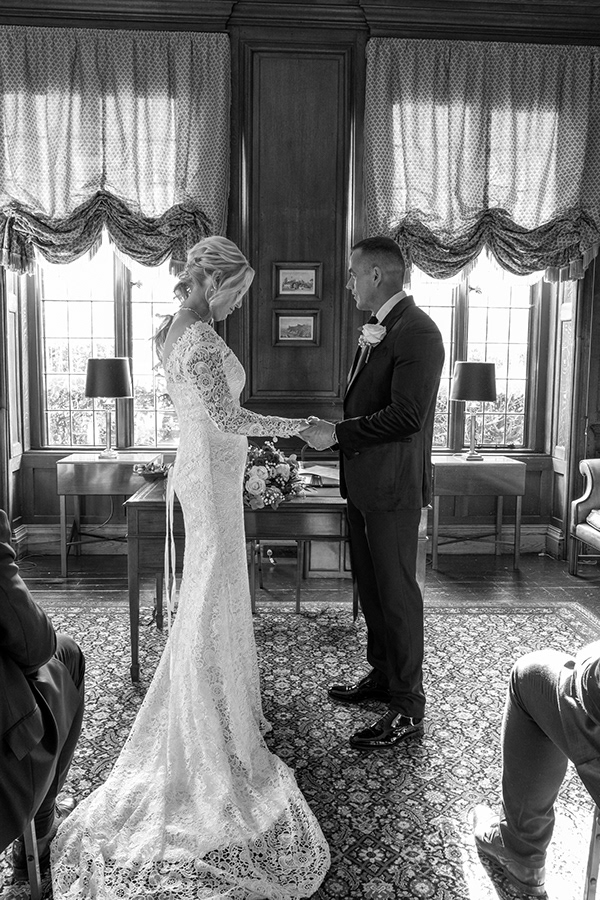 Black and white photograph of a bride and groom standing together during an intimate indoor wedding ceremony.