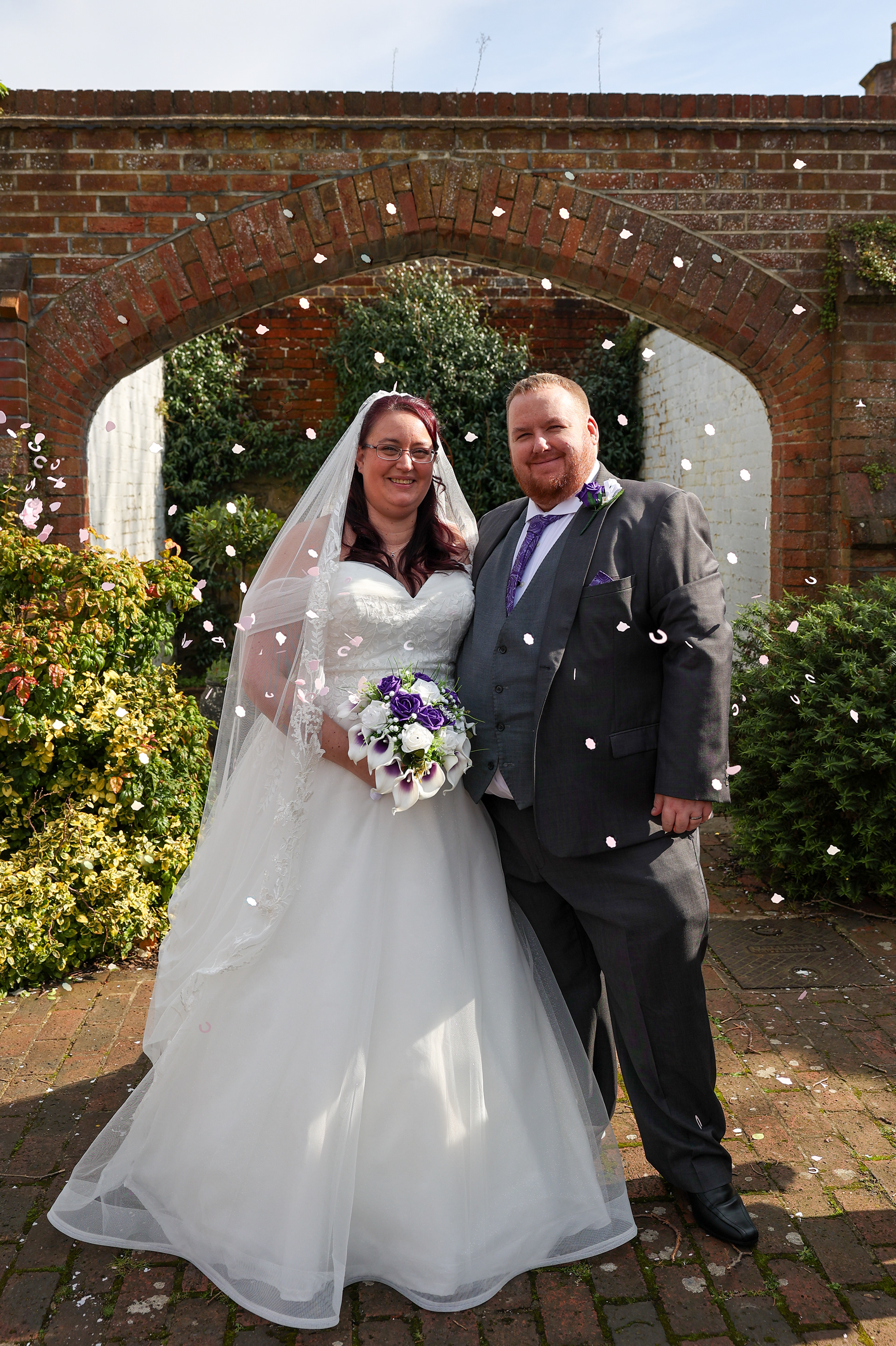 Bride and groom posing beneath a brick archway while confetti falls around them after the wedding ceremony.