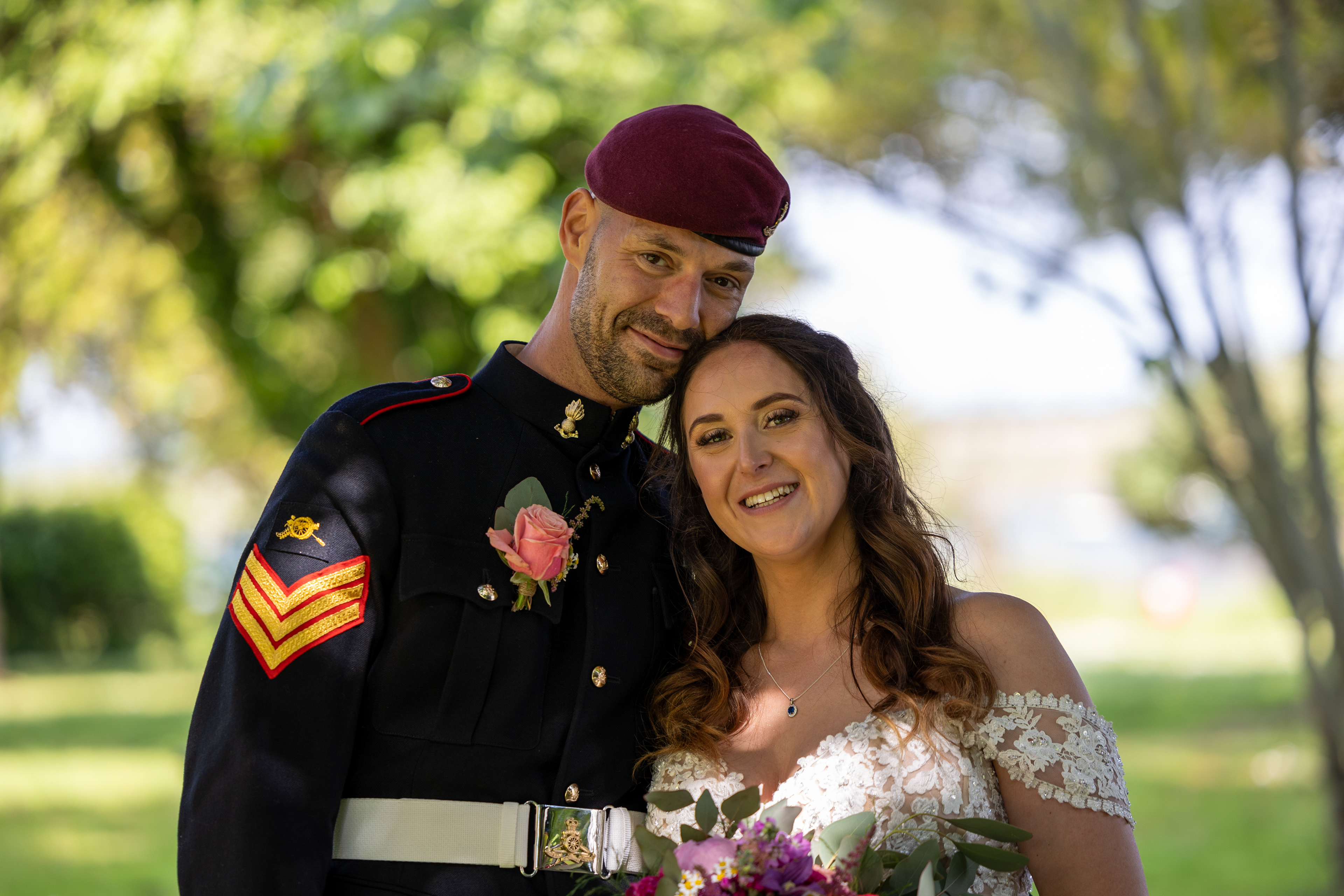 Bride and groom smiling together outdoors beneath trees, with the groom wearing formal military uniform and the bride holding a bouquet.