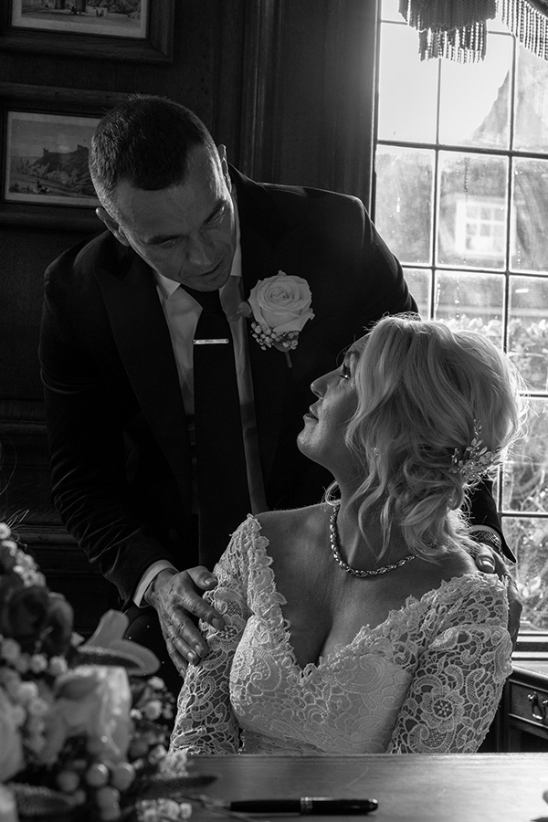 Black and white photograph of a bride and groom sharing an intimate moment indoors beside a window during their wedding ceremony.