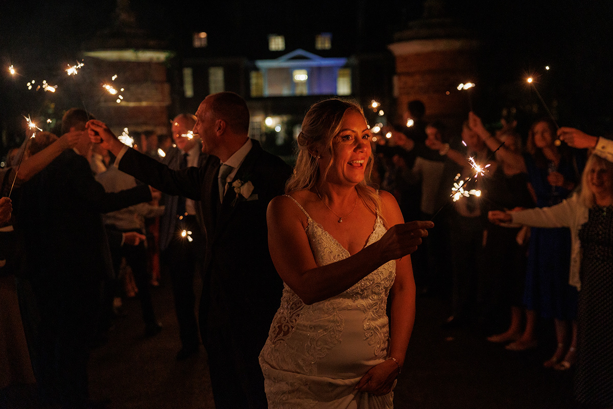 Bride and groom holding sparklers during an evening wedding celebration surrounded by guests outside a country house venue at night.