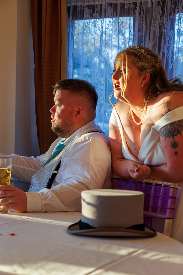Bride and groom seated together by a window during a wedding reception, illuminated by warm evening light.