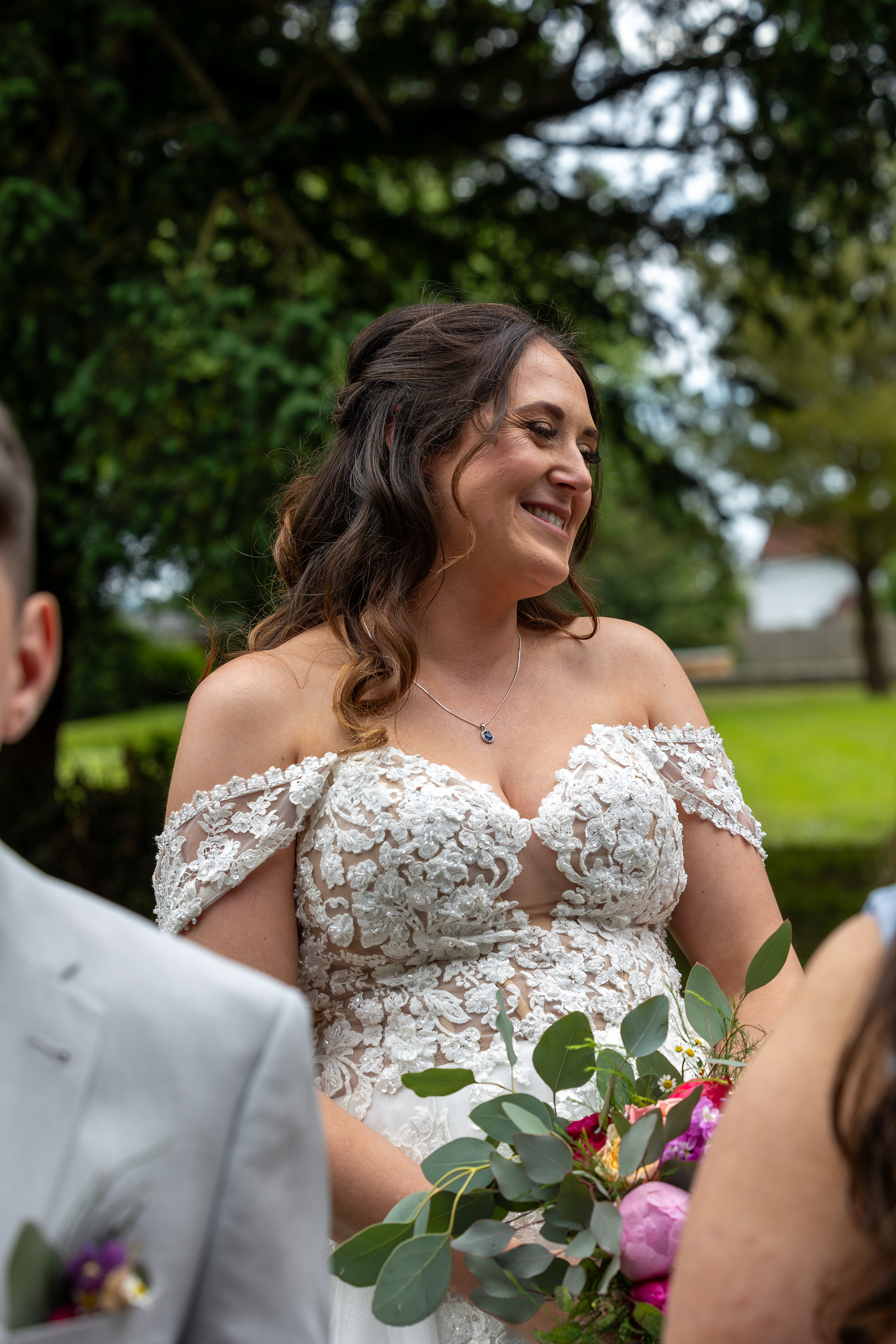 Bride in a lace wedding dress smiling during an outdoor wedding ceremony while holding a colourful bouquet.