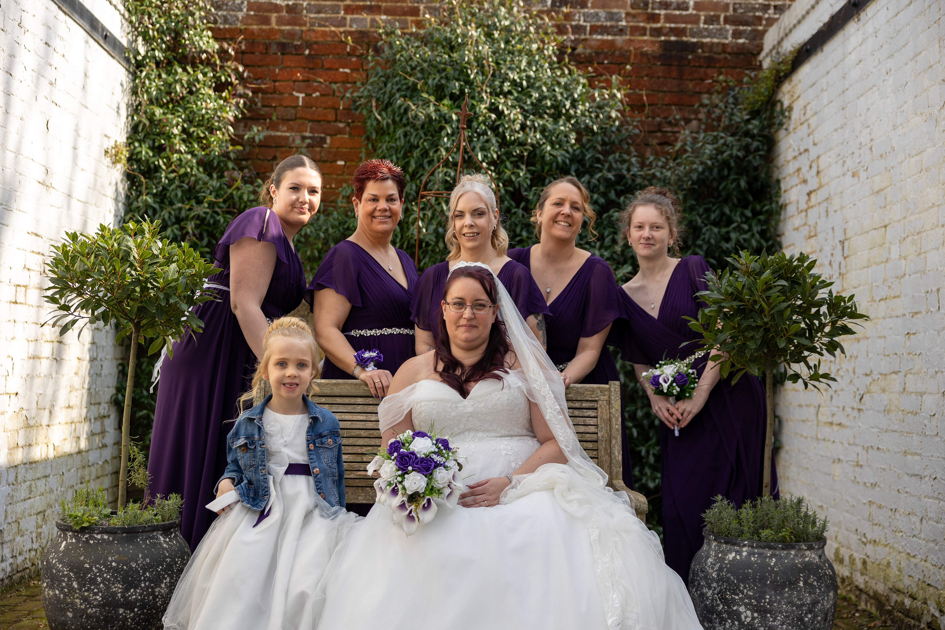 Bride seated with bridesmaids in purple dresses and a flower girl during a formal outdoor wedding group portrait.