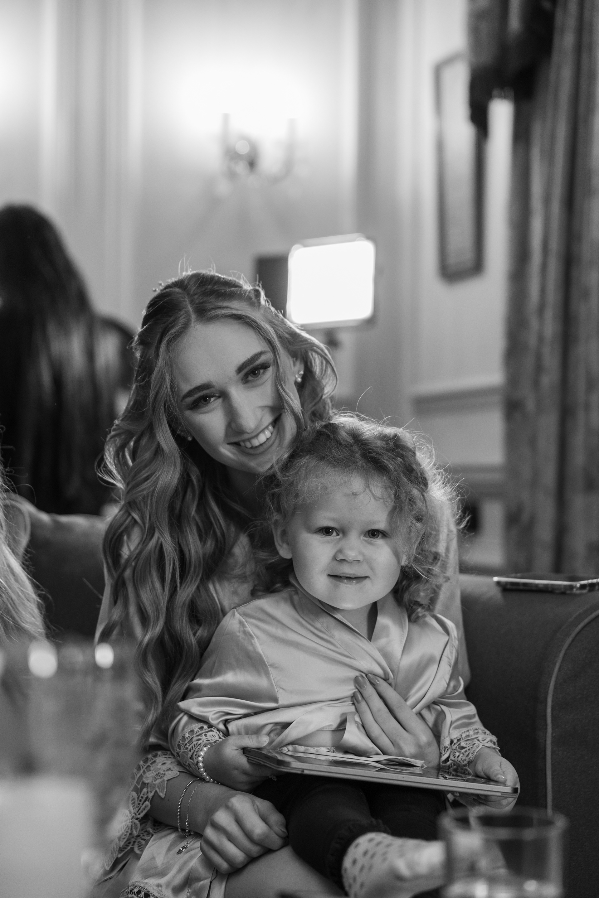 Black and white photograph of a smiling bridesmaid sitting with a young flower girl during wedding preparations indoors.