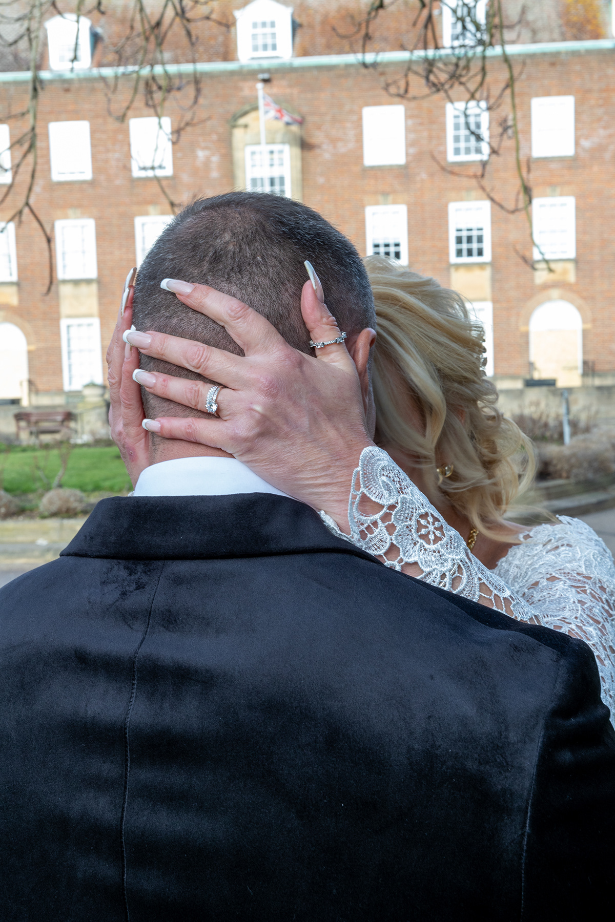Close-up photograph of a bride embracing the groom outdoors, with wedding rings and lace sleeve details visible.