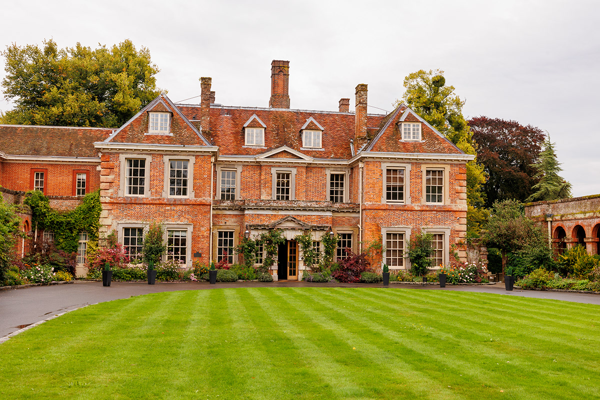 Large red brick manor house wedding venue with landscaped gardens and striped lawn under an overcast sky.
