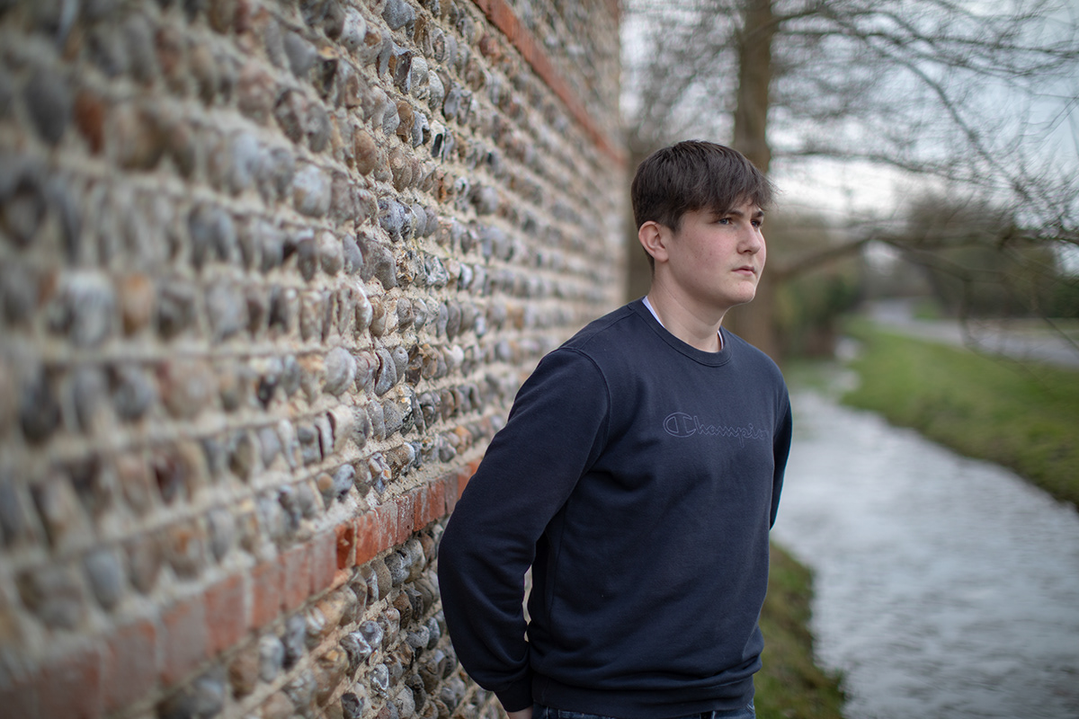 Teenage boy standing beside a textured stone wall near a riverside path during an outdoor portrait session.