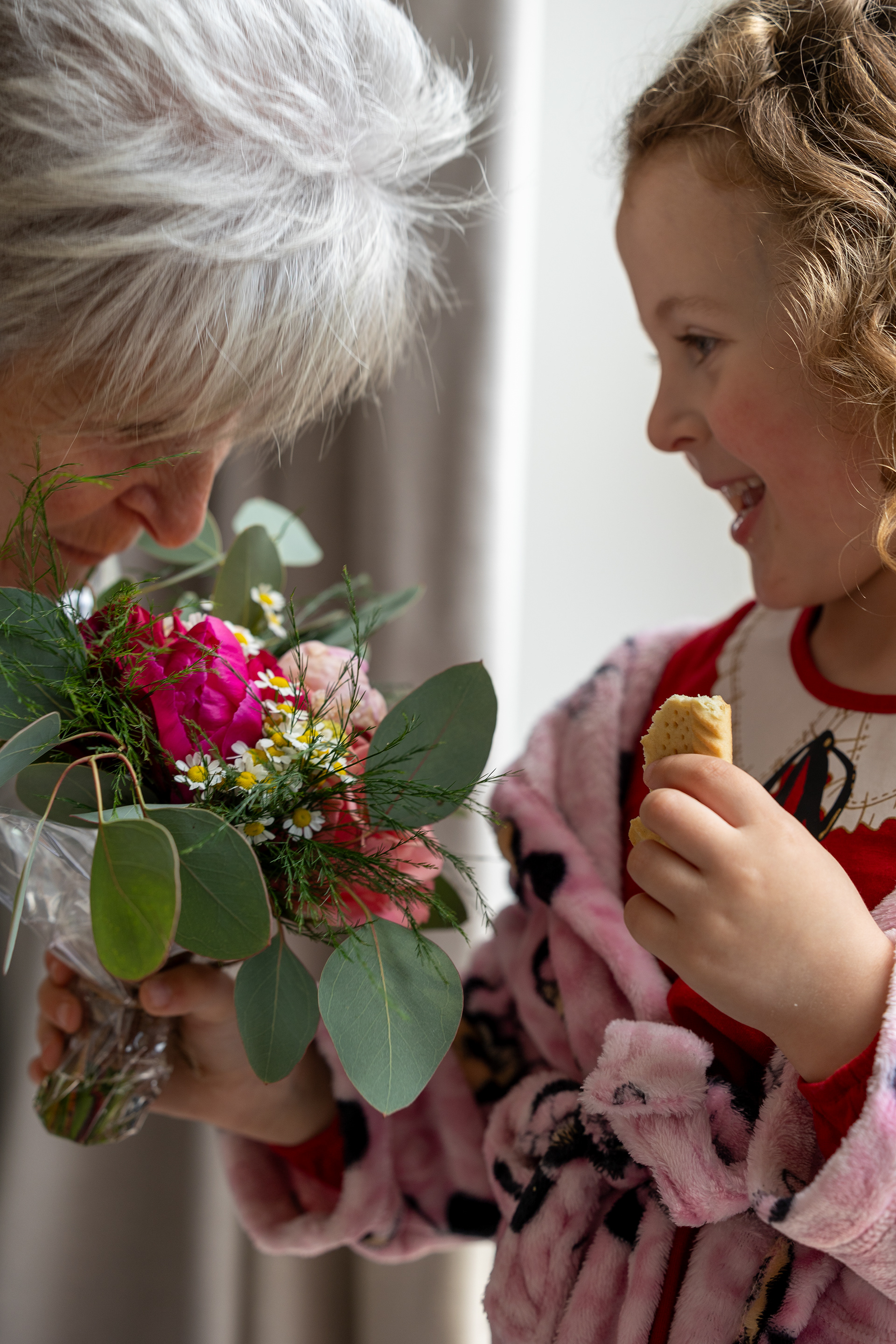 Young flower girl smiling while holding a biscuit beside an older woman holding a colourful wedding bouquet indoors.