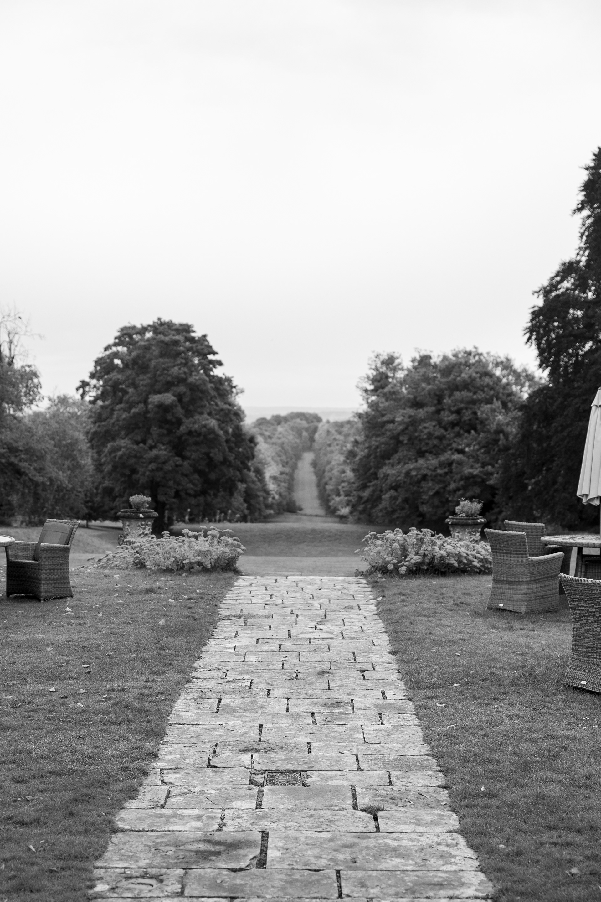 Black and white photograph of a stone pathway leading through landscaped gardens toward distant trees at a countryside wedding venue.