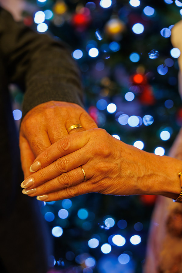 Close-up photograph of an older couple holding hands with wedding rings visible, set against softly blurred Christmas tree lights and festive bokeh.