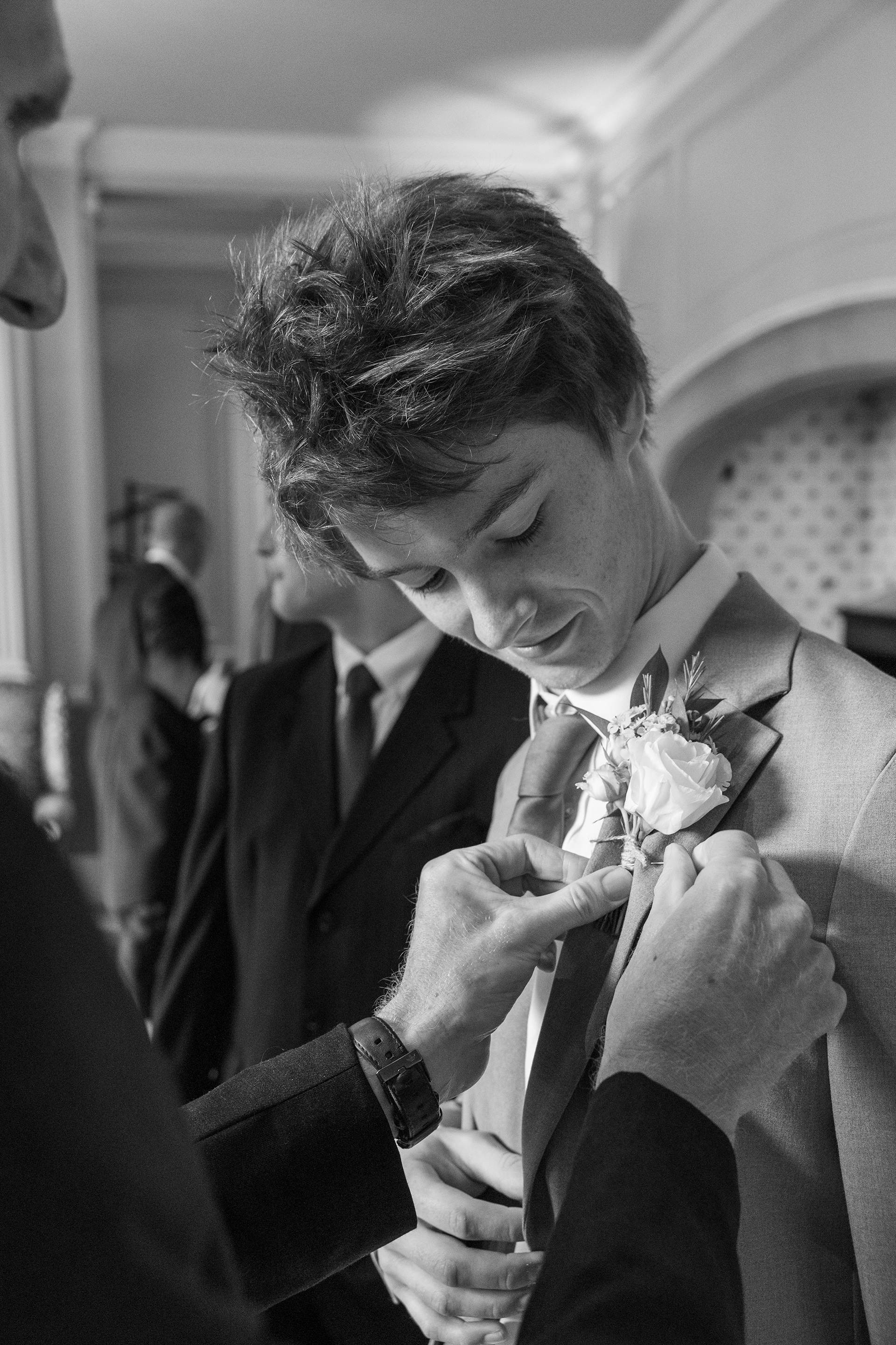 Black and white photograph of a groom having his buttonhole adjusted while getting ready before a wedding ceremony.