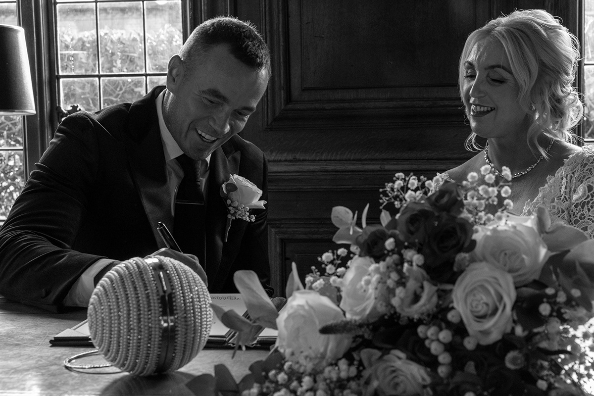 Black and white photograph of a groom signing the wedding register while the bride smiles beside him indoors.