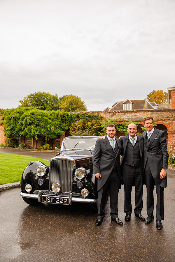 Three men in formal wedding attire standing beside a vintage black wedding car outside a countryside venue on an overcast day.
