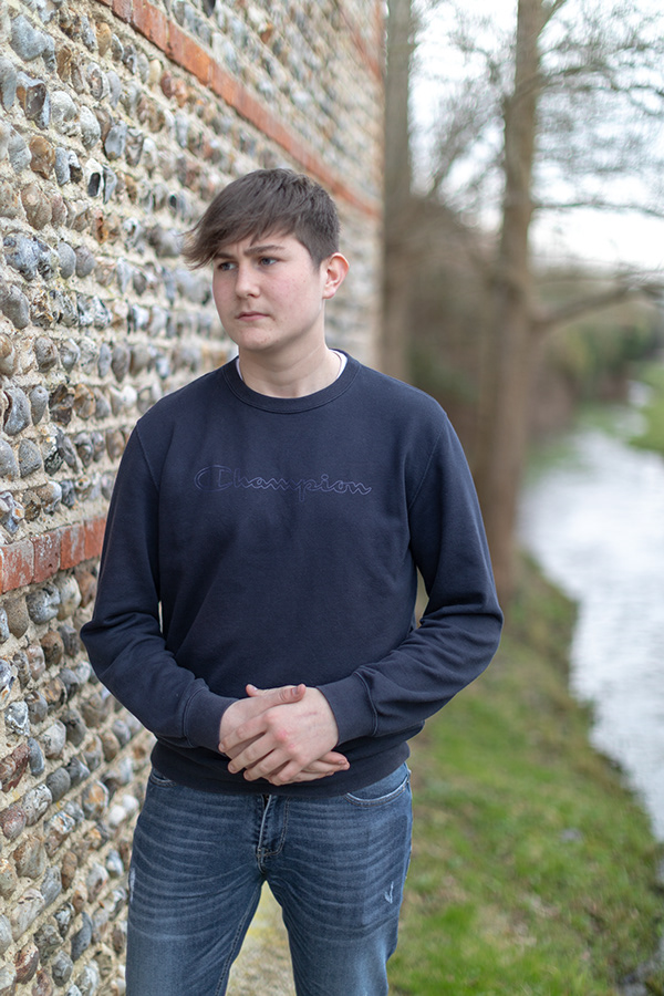 Teenage boy standing beside a textured stone wall near a riverside path during an outdoor portrait session.