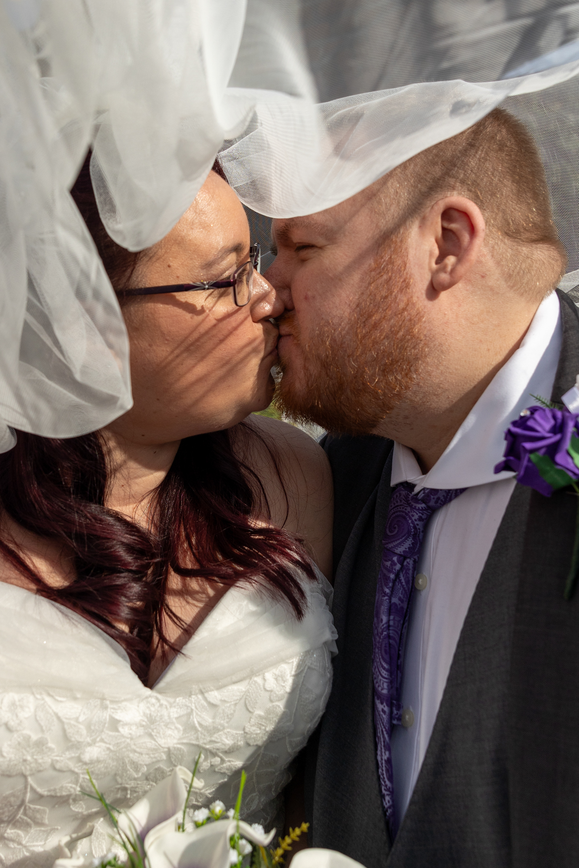Close-up photograph of a bride and groom kissing beneath a wedding veil during an outdoor portrait session.