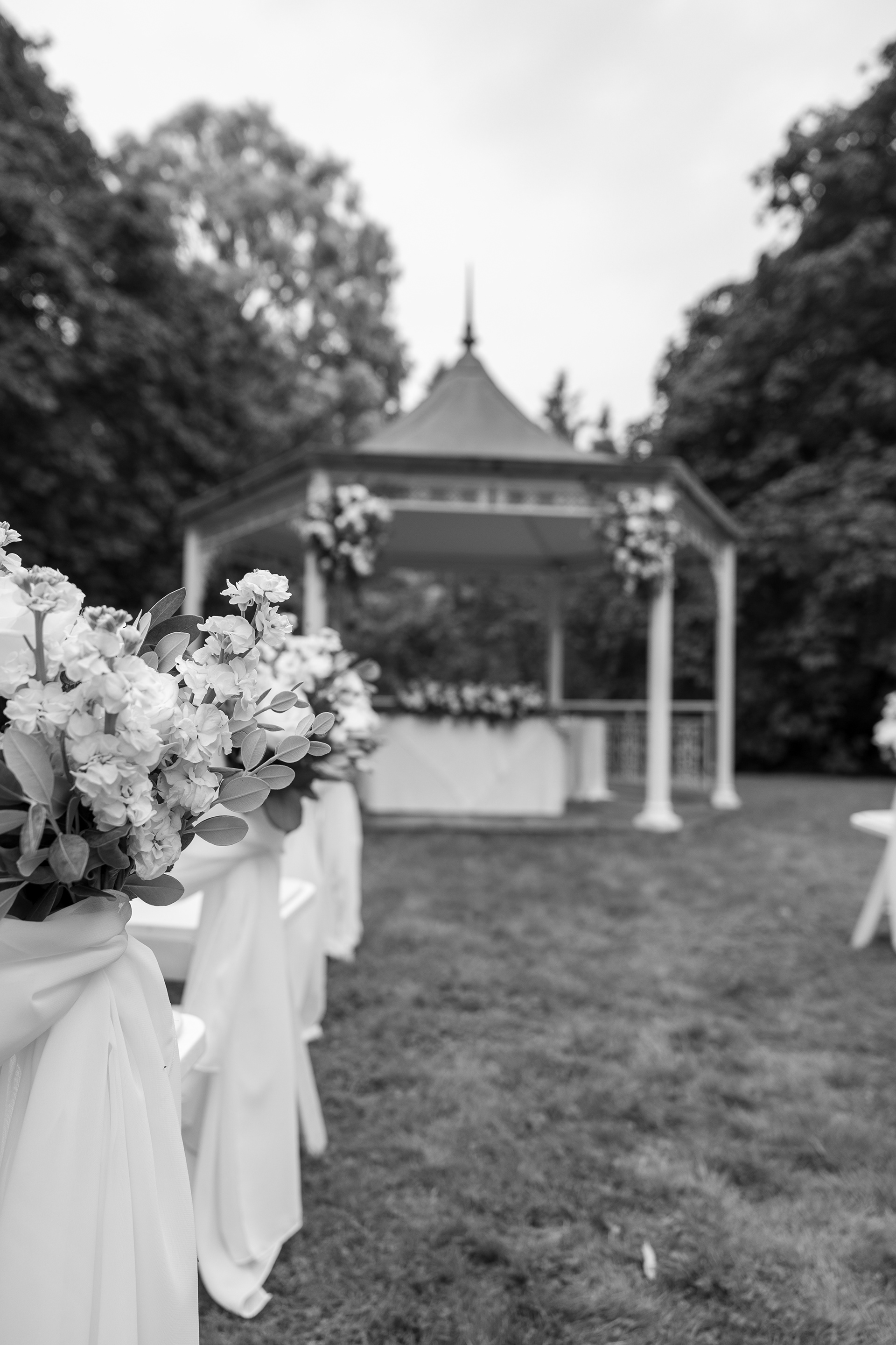 Black and white photograph of an outdoor wedding ceremony area with floral aisle decorations leading toward a white gazebo surrounded by trees.