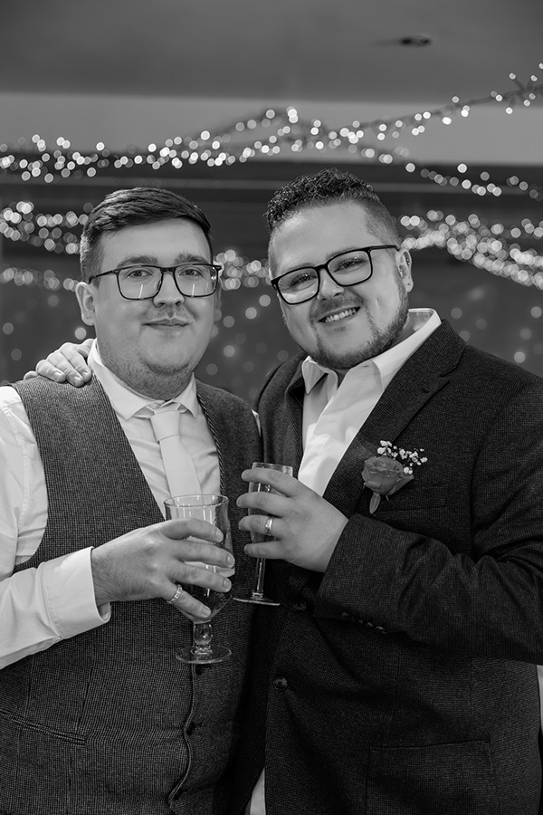 Black and white wedding portrait of two men smiling together while holding drinks, surrounded by soft fairy light bokeh at an evening reception.