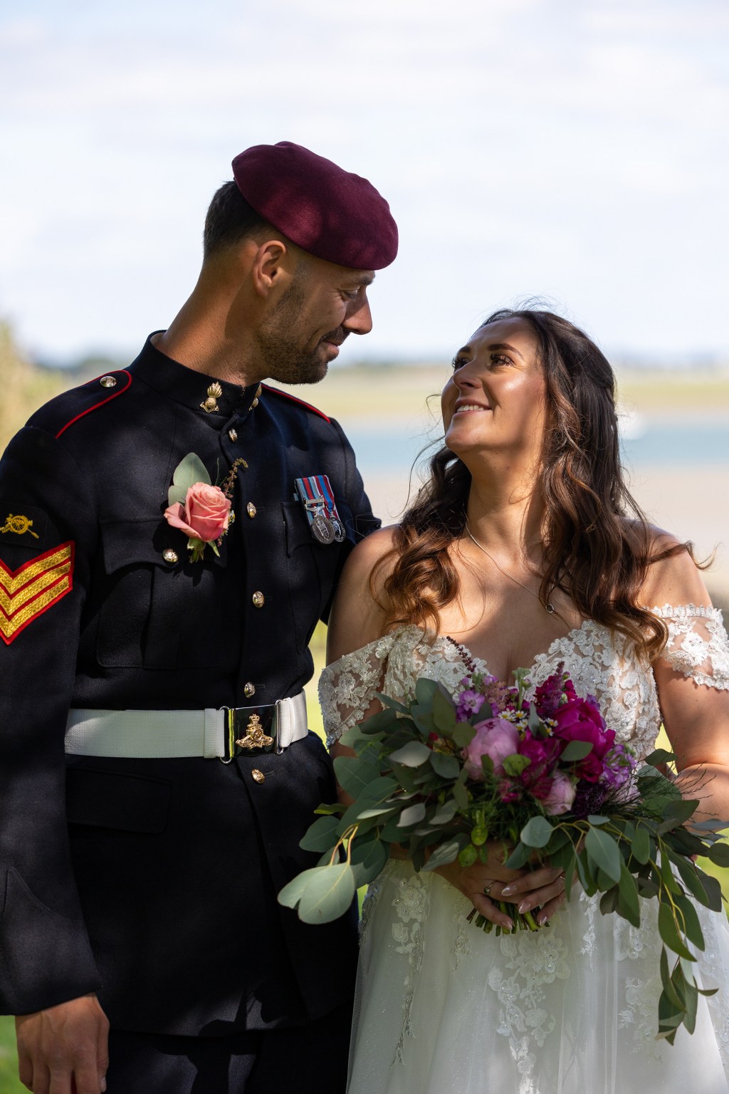 Bride and groom looking at each other outdoors near the coast, with the groom wearing formal military uniform and the bride holding a bouquet.