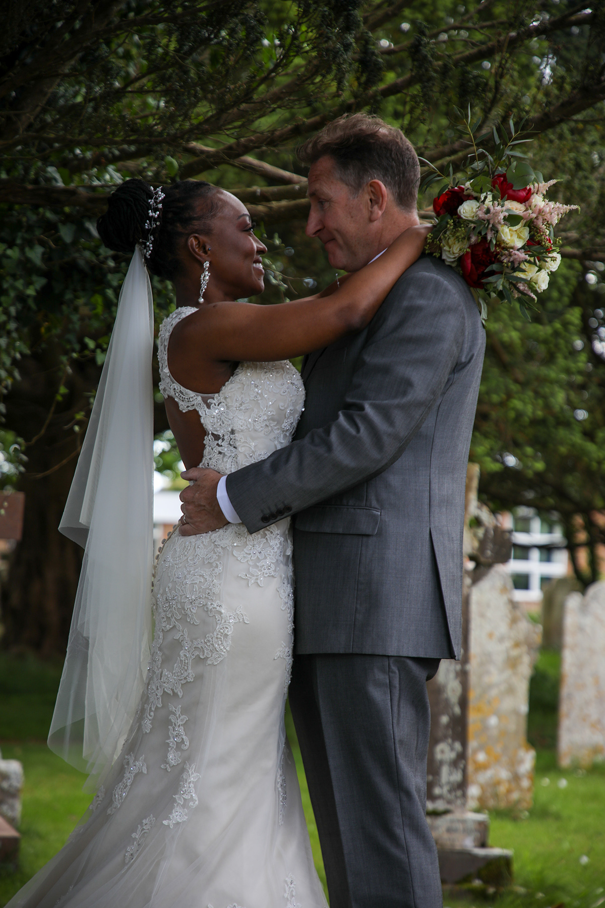 Bride and groom embracing beneath trees outdoors after their wedding ceremony, with the bride holding a bouquet behind the groom’s shoulders.
