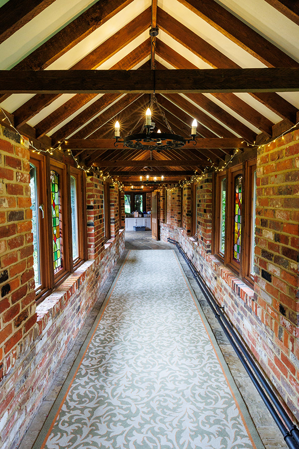 Long brick corridor with exposed wooden beams, hanging chandeliers, stained glass windows, and warm fairy lights inside a rustic wedding venue.